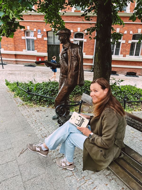 Astrid Engel sitzt auf der Bronze-Bank vor der Bronzestatue von Brunon Zwarra und hält sein Buch "Die Danziger" in der Hand.